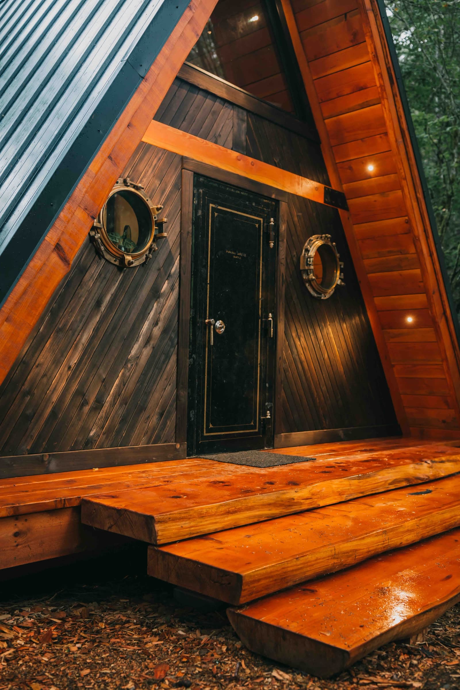 Close view of the A-frame’s dark-stained cedar gable, with two round brass portholes either side of a heavy black door.