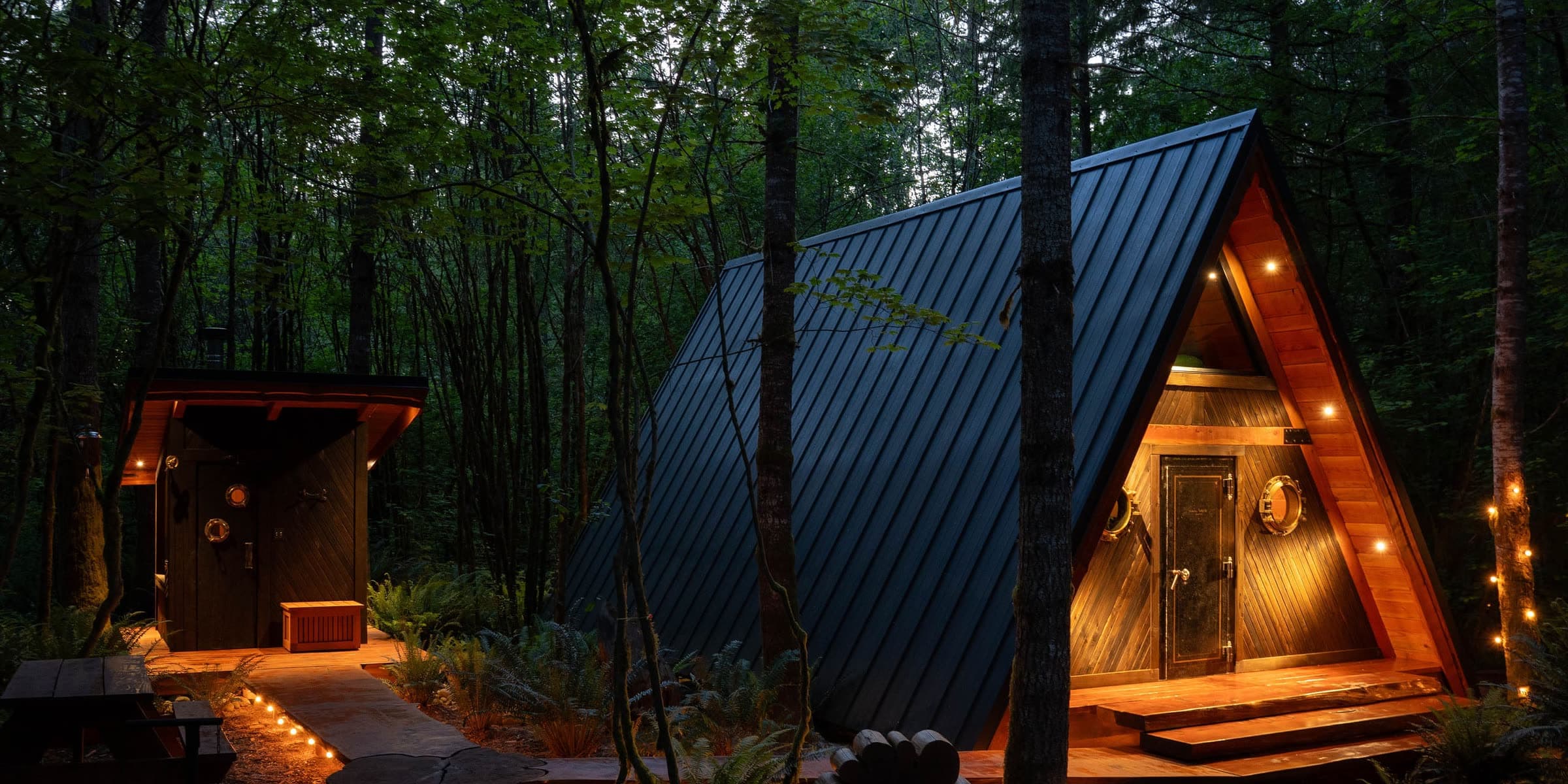 The A-frame cabin and its cedar sauna at dusk, warm light glowing in the gable and along a wooden boardwalk, surrounded by tall forest.