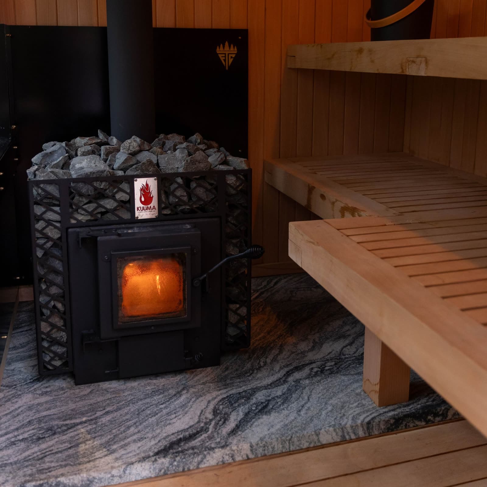 The wood-burning stove glowing inside the cedar sauna, with stacked stones on top and pale cedar benches beside it.