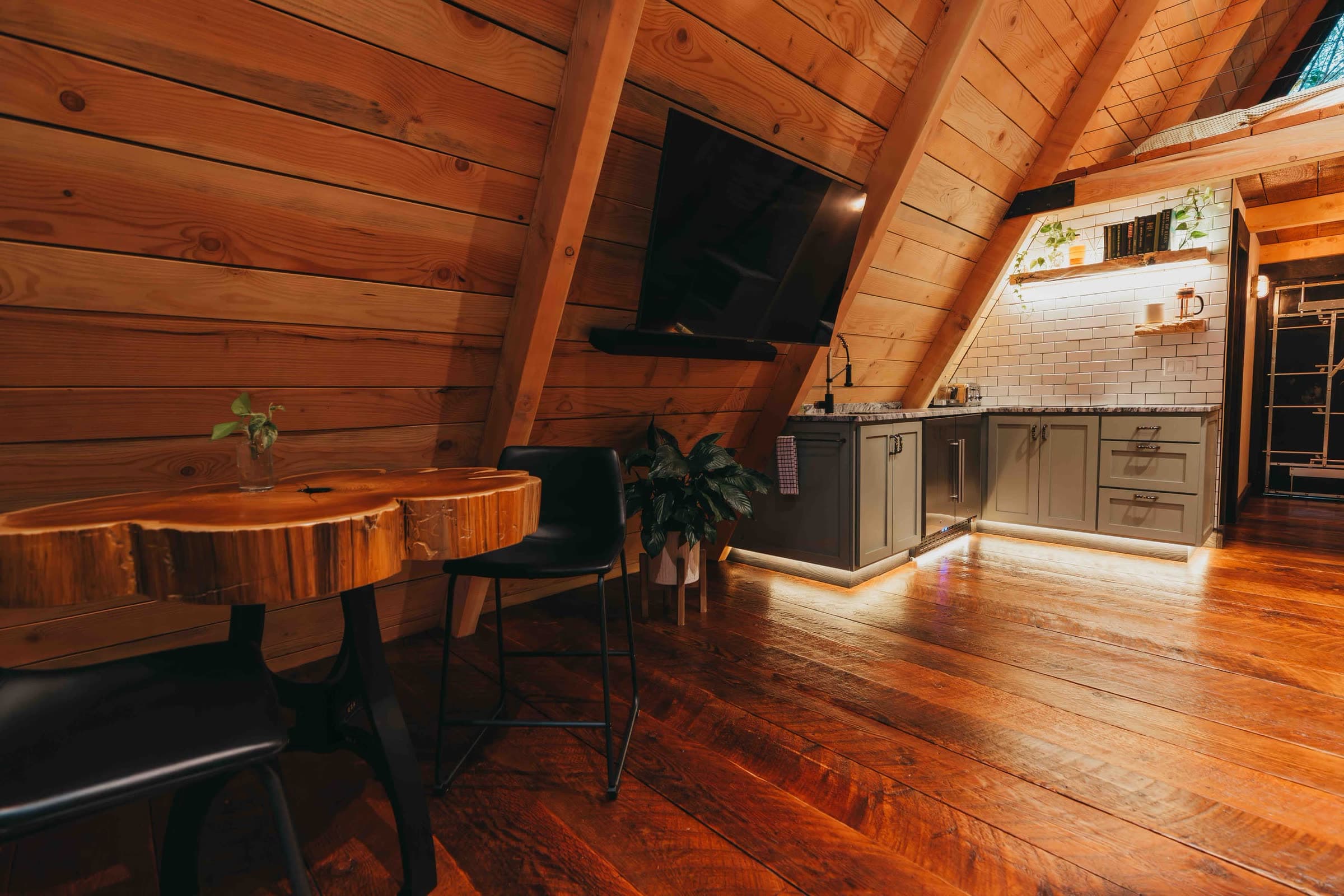Inside the cabin: a live-edge slab dining table, a wall-mounted TV and a small kitchen with sage-green cabinets and warm under-cabinet lighting, on wide plank floors beneath a cedar ceiling.