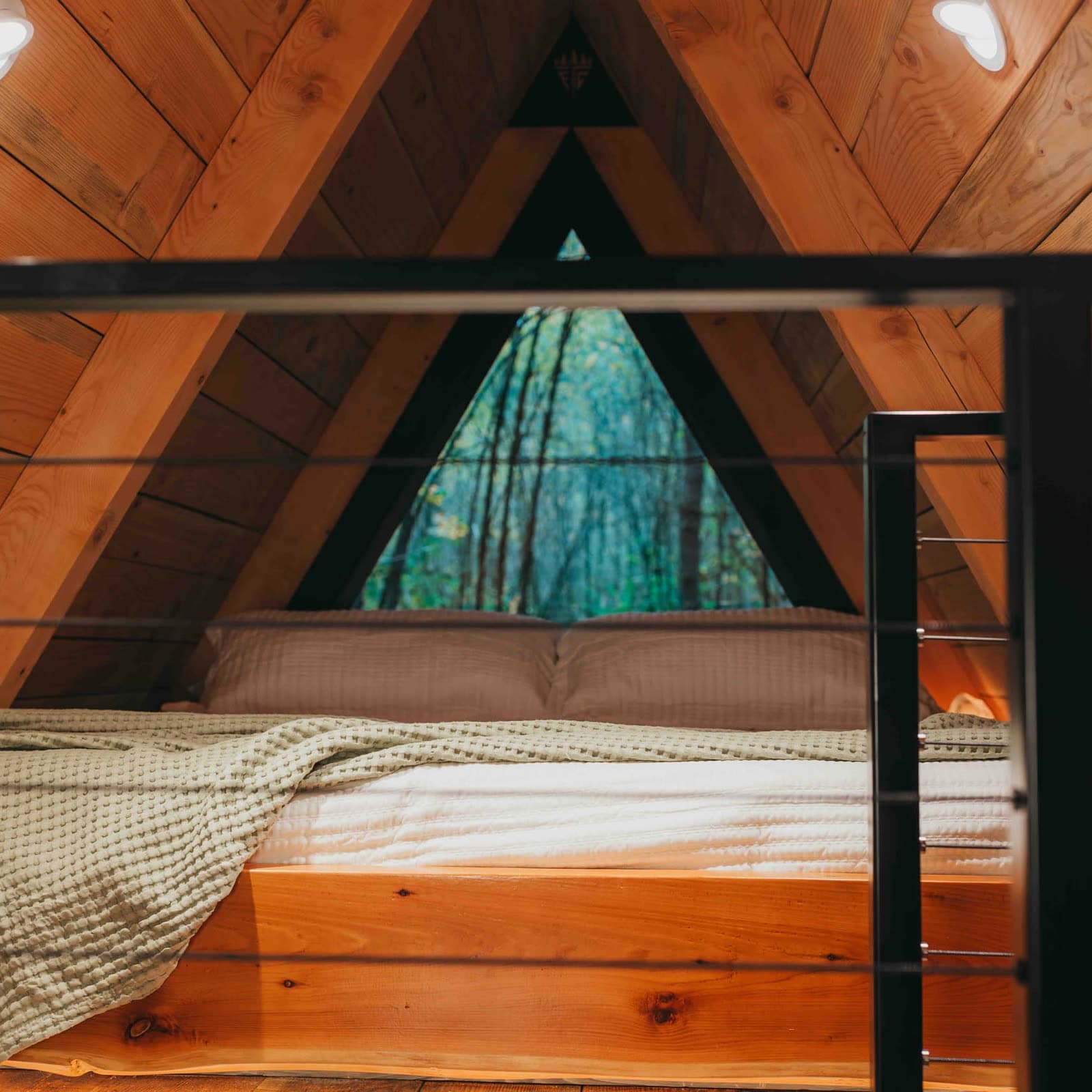 The loft bed beneath the cabin’s triangular glass gable wall, with a live-edge cedar footboard, black cable railing and forest visible through the glass.