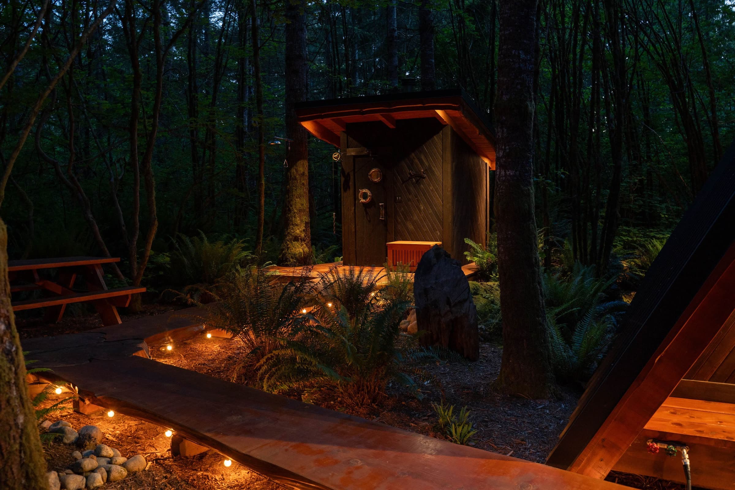 A lit live-edge boardwalk leading past a standing stone toward the cedar sauna at dusk, with a picnic table and ferns in the dark forest.
