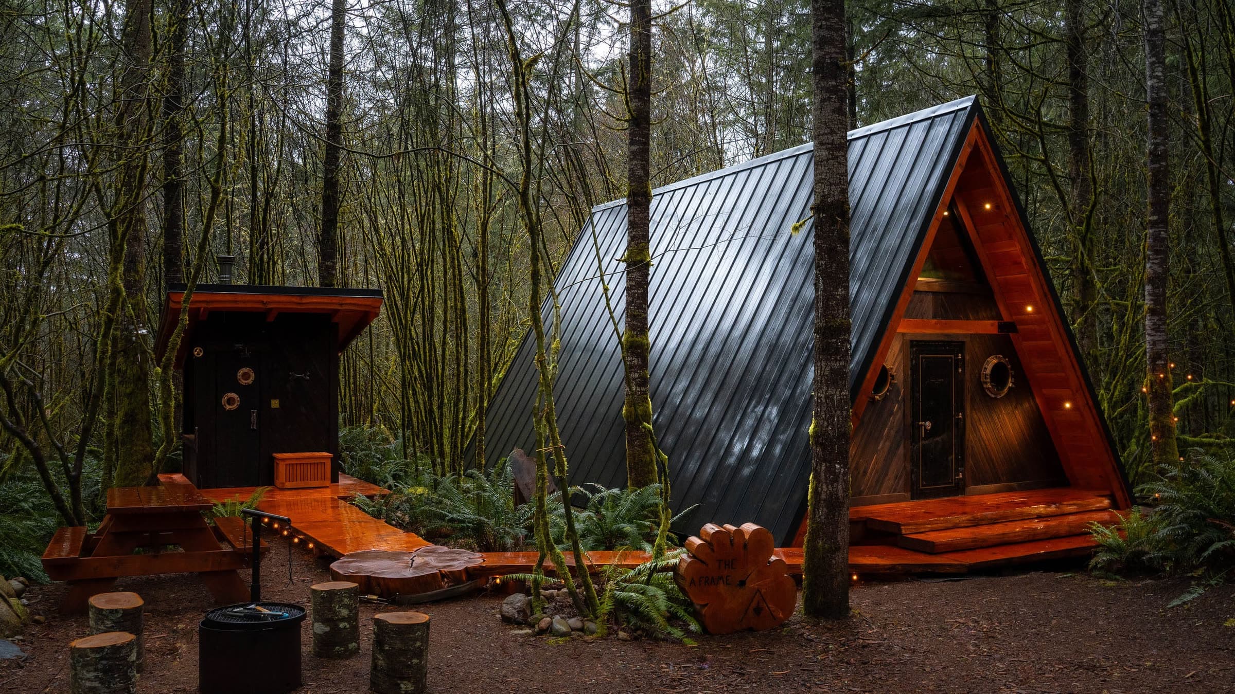 The A-frame cabin at twilight in the forest, its dark steel roof and cedar gable lit by small warm lights, with the cedar sauna cabin to the left and a round log-slab sign beside the boardwalk.