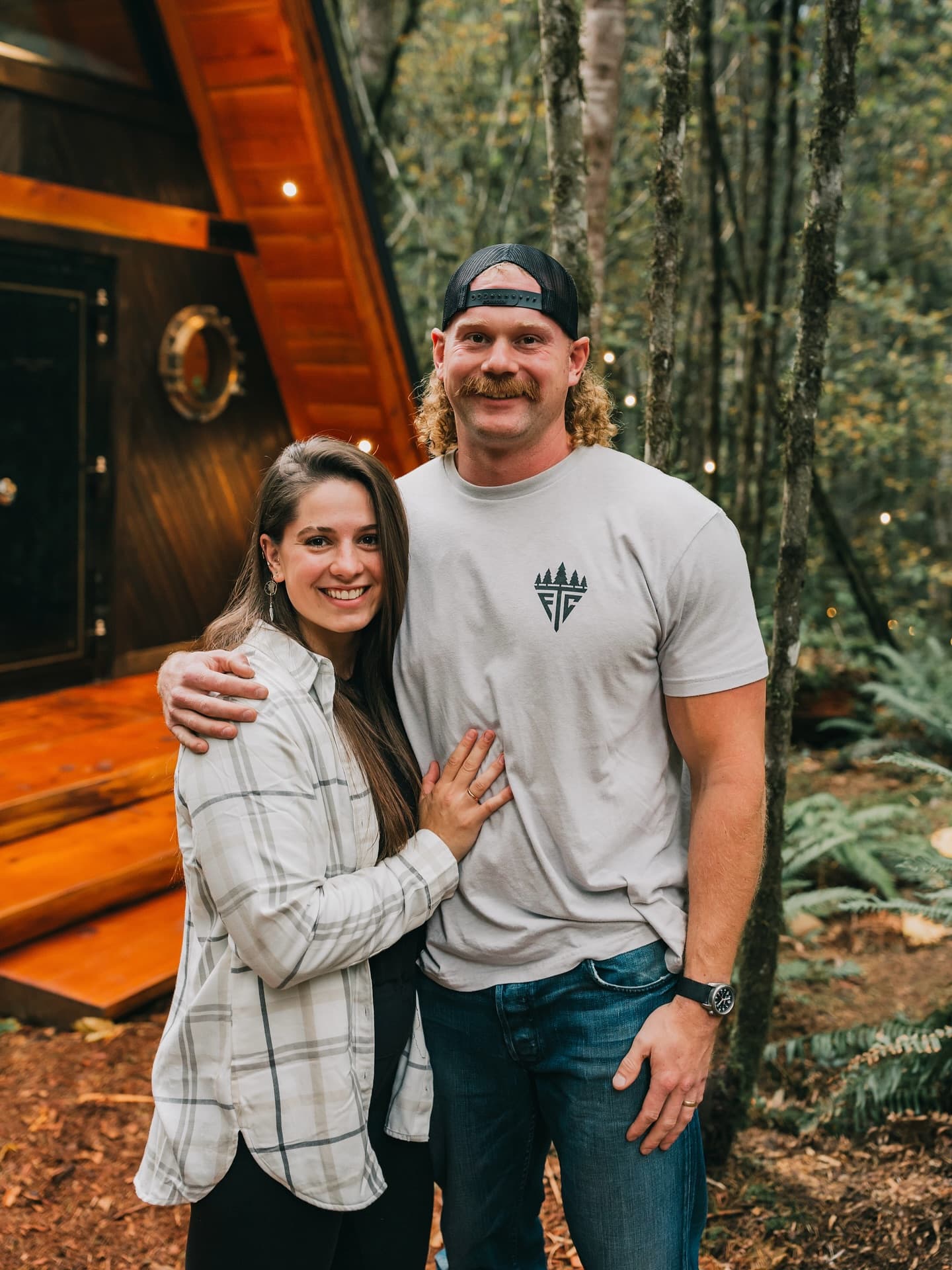 Katie and Ryan Fromm, arm in arm in front of the A-Frame’s lit cedar gable, with ferns and string lights behind them.