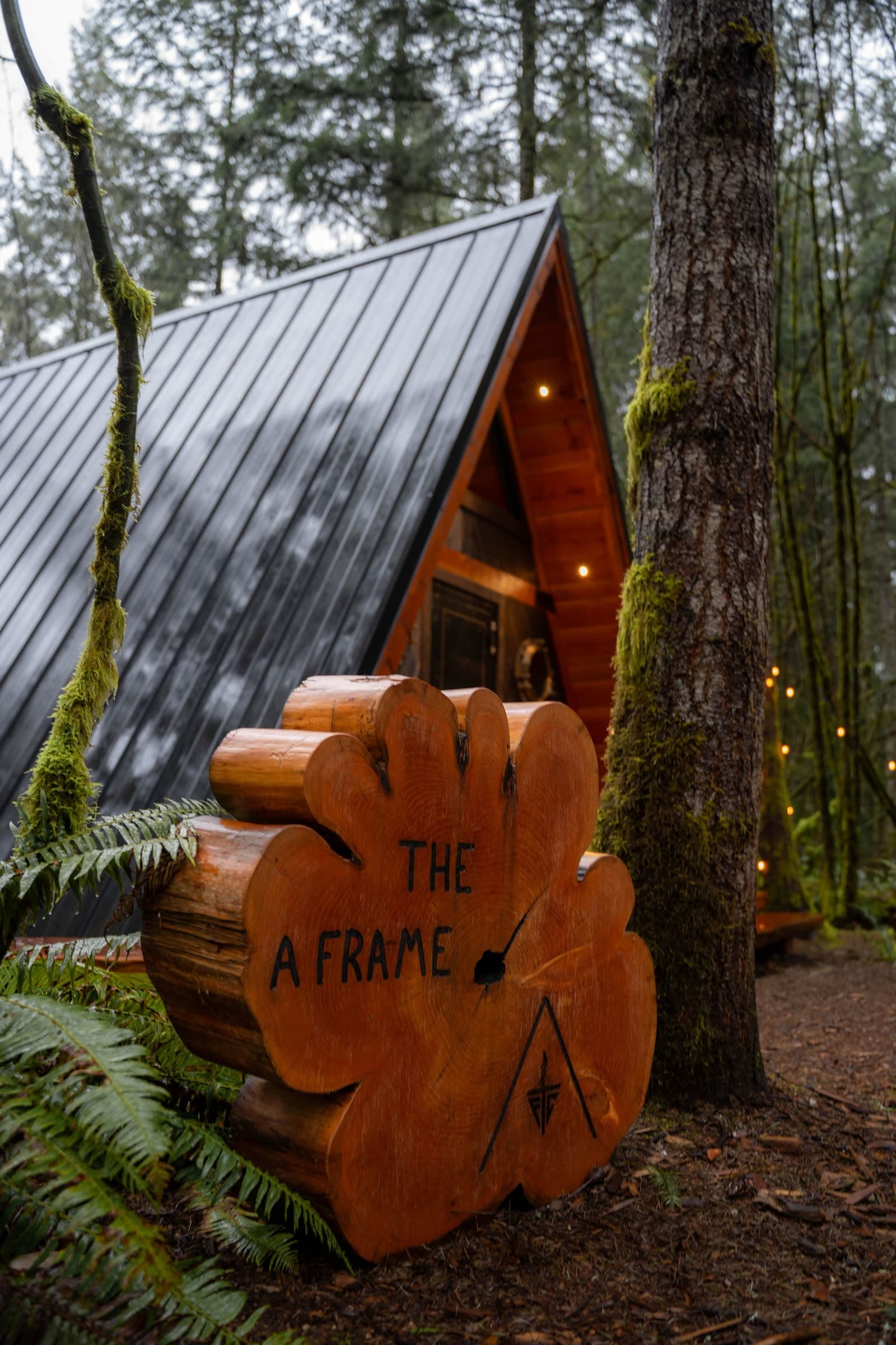 A cedar log-slab sign reading “THE A FRAME” in front of the cabin’s steel roof, among ferns and moss.